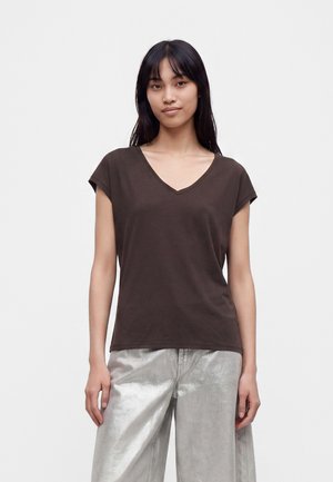 Jeune femme aux longs cheveux foncés portant un T-shirt ample marron foncé à col en V et un pantalon large argenté brillant, debout devant un fond uni.