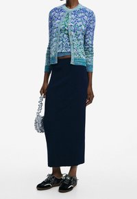 Sinivihreä kuvioitu neulejakku vastaavan kuosin lyhyen yläosan päällä, yhdistetty tumman ribbineuloksiseen hameeseen ja mustiin nauhallisiin lenkkareihin. Pieni teksturoitu laukku.