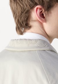 Lys grå jakke med en strukturert krage som har preget tekst. Glatt stoff og enkel design, vist fra baksiden med en hvit skjorte.