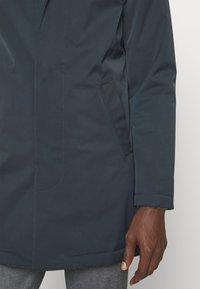 Donkerblauwe waterdichte jas met een strakke uitstraling, rechte snit en zijzakken. Gemaakt van soepel, duurzaam materiaal met minimale stiksels.