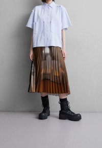 Jasnoniebieska koszula krótki rękaw z dwiema kieszeniami, zestawiona z brązową spódnicą plisowaną z gradientowym wzorem i czarnymi, masywnymi butami na sznurówki.