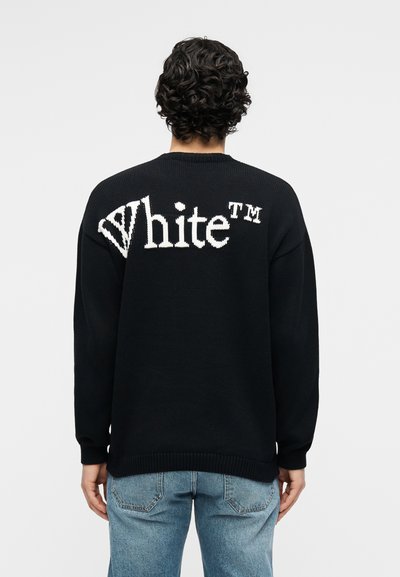 Άτομο με σγουρά μαλλιά που φοράει μαύρο πουλόβερ με τη λέξη "White™" σε μεγάλα λευκά γράμματα στην πλάτη, συνδυασμένο με μπλε τζιν.