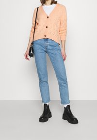 Růžová pletená cardigan s vzorem šípů, zapínání na knoflíky, bílý tričko pod tím, spárované se světle modrými rovnými džíny a černými platformovými botami.
