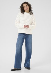 Witte gebreide trui met een geribbelde structuur en open patroon, gecombineerd met wijde blauwe jeans en zwarte platte schoenen.