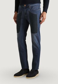 Jean en denim de couleur bleu foncé, avec des panneaux en tissu noir sur les cuisses, design droit et configuration classique à cinq poches.