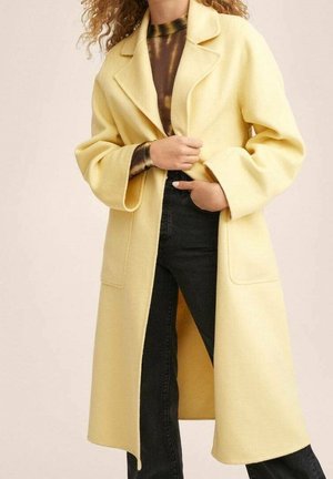 Kvinde iført en lang, lysegul frakke over en sort og brun batikfarvet skjorte og sorte bukser, med den ene hånd i frakkens lomme.