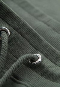 Dunkelgrüner, gerippter Stoff mit einem Metall-Ösen und einem passenden Kordelzug, der die Textur und ein sportliches Design betont.