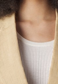 Beżowa lniana marynarka z teksturowanymi krawędziami tkaniny, nałożona na jasnoszary prążkowany top na ramiączkach. Zaokrąglony dekolt i subtelny kontrast kolorów widoczny.