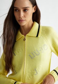 Maglione giallo con zip e colletto, caratterizzato da strisce nere, logo "LIU JO" glitterato, lavorazione a maglia strutturata e polsini a costine.