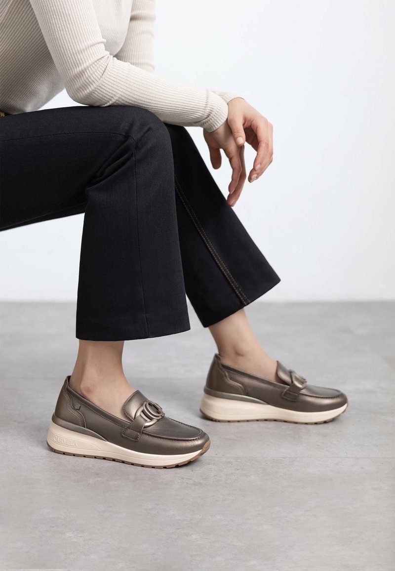 Zapatos slip-on de cuero bronce con un detalle de hebilla redonda, montados sobre una suela de goma crema, combinados con jeans oscuros y un suéter claro.