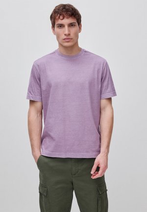 Joven hombre con camiseta de manga corta de color lila claro y pantalones cargo verde oliva, de pie frente a un fondo claro y liso.