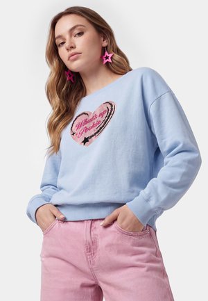 Kvinde med langt hår, der bærer lyserøde stjerne-øreringe, lyseblå sweatshirt med hjerte og tekst, og lyserøde bukser med hænderne i lommerne.