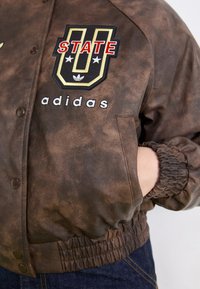 Rjava bomberska jakna z mehkimi, teksturiranimi površinami. Ima vezeno oznako "STATE U" in besedilo "adidas" na prsih, elastične manšete in zaklopne gumbe.