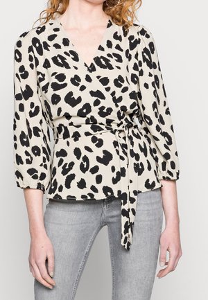 Vrouw die een beige wikkelblouse draagt met zwarte luipaardvlekken en driekwartmouwen, gecombineerd met lichtgrijze spijkerbroek.