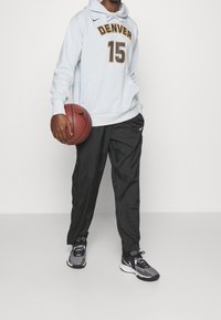 Persona con pantalones negros y zapatillas grises sosteniendo un balón de baloncesto, vistiendo una sudadera de Denver azul claro con el número 15.