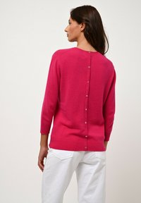 Suéter fucsia con una textura suave, escote en V, mangas de tres cuartos y una fila de botones en la parte posterior. Combinado con pantalones blancos.