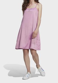 Różowa sukienka bez rękawów w luźnym kształcie A, z małym logo na klatce piersiowej i gładką, błyszczącą teksturą. Zestawiona z białymi sneakersami.