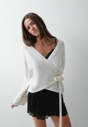 Femme aux longs cheveux bruns portant un pull cache-cœur blanc sur une robe noire bordée de dentelle, debout devant un fond blanc uni.