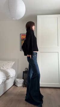 Άτομο με μαύρο πουλόβερ και τζιν με φαρδιά μπατζάκια στέκεται πλαγίως σε ένα υπνοδωμάτιο με λευκό κρεβάτι, τουαλέτα, φωτιστικό και κορνιζαρισμένη αφίσα.