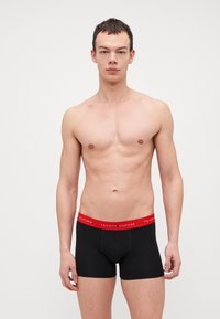 Schwarze Boxer-Briefs mit einem roten Gummibund, auf dem das Logo "TOMMY HILFIGER" zu sehen ist. Glattes Material mit enger Passform und ohne zusätzliche Muster oder Akzente.