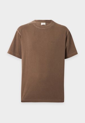Mann steht, trägt ein braunes kurzärmeliges T-Shirt, beige weit geschnittene Hose und schwarze Schuhe, eine Hand in der Tasche.