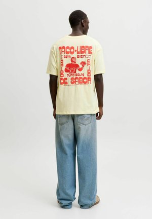 Homme portant un t-shirt jaune pâle avec un grand graphisme rouge et un texte sur les tacos, associé à un jean large bleu clair et des chaussures beige, vu de dos.