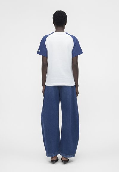 Άτομο στέκεται με την πλάτη, φοράει λευκό και μπλε κοντομάνικο πουκάμισο, φαρδιά μπλε τζιν και μαύρα ψηλοτάκουνα παπούτσια σε απλό φόντο.