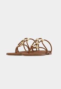 Καφέ επίπεδα σανδάλια με πολλαπλά λεπτά λουριά και μπεζ πλεκτές λεπτομέρειες, σχεδιασμός με ανοιχτά δάχτυλα και χαμηλό τακούνι πάνω σε απλό φόντο.
