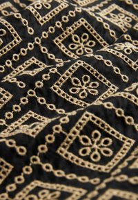 Černá tkanina s béžovými vyšívanými geometrickými a květinovými vzory tvořícími diamantové tvary a propojenými smyčkami.