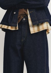 Jakna in hlače iz temno modrega jeansa z rumenim šivanjem. Plastičasta karirana srajca z bež in rumenimi odtenki, vidna na zaključku. Kovinski gumbi.