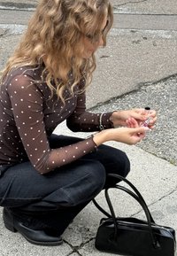 Model wears a sheer brown polka-dot top and black jeans, holding a nail polish bottle. A small black handbag rests beside her on the ground.
