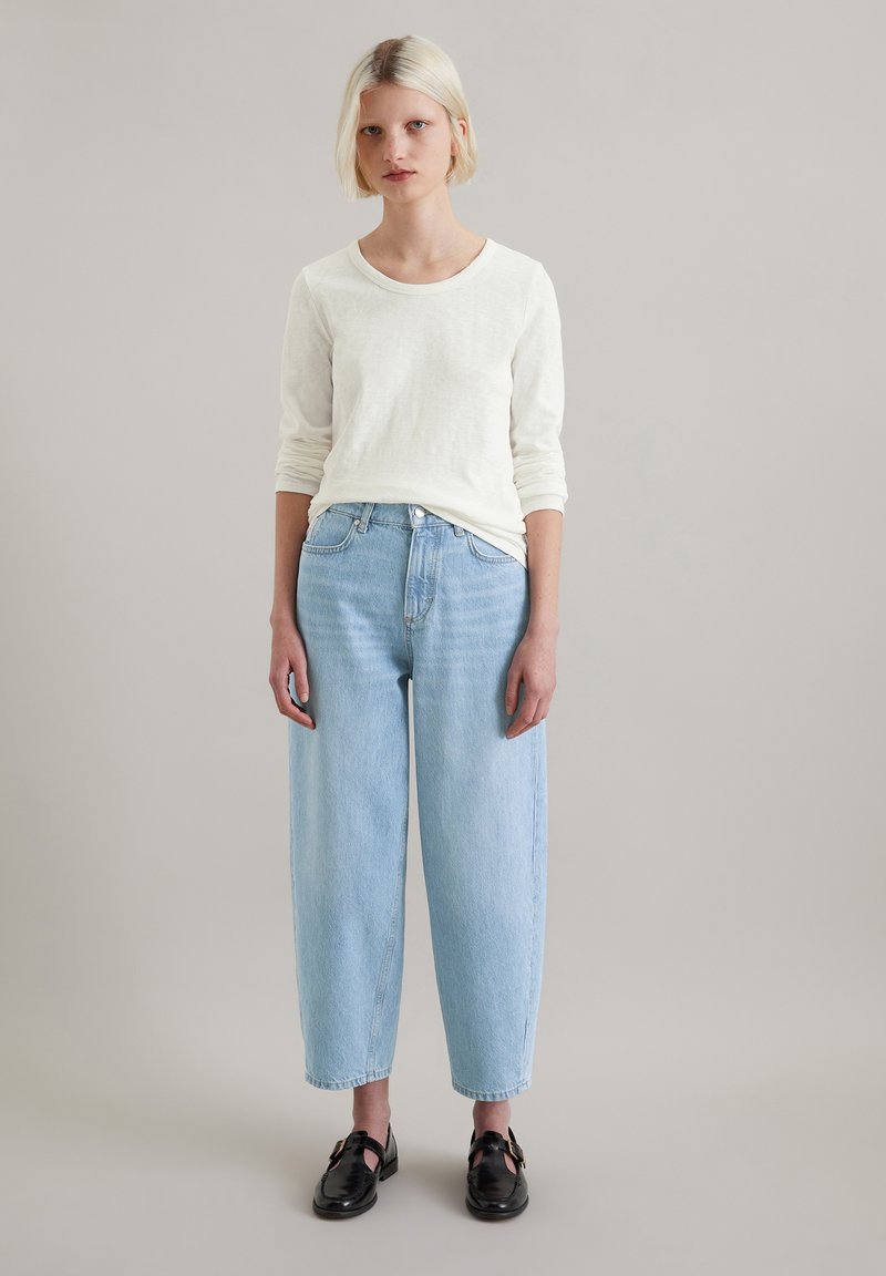 Biała bawełniana koszula z długim rękawem zestawiona z jasnoniebieskimi, luźnymi dżinsami i czarnymi skórzanymi butami z paskami.