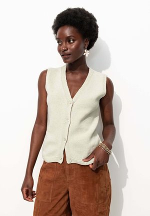 Cardigan sans manches vert clair avec un col en V et des boutons sur le devant. Porté par-dessus un pantalon en suédé marron. Tricot simple avec texture. Aucun motif ni accent visible.