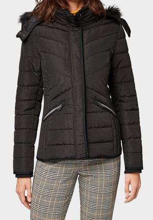 Kobieta w czarnej pikowanej kurtce z kapturem obszytym futrem i kieszeniami na zamek, w połączeniu z spodniami w kratę.