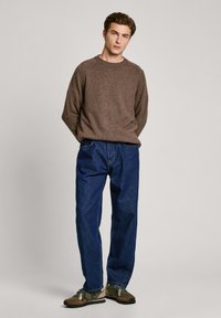 Jersey de punto marrón, jeans de mezclilla azul holgados y zapatillas verdes/negras con acentos marrones. El modelo está de pie contra un fondo claro.