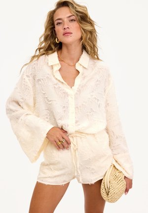 Femme portant une blouse crème texturée à boutons et un short assorti, tenant une pochette en paille tissée, accessoirisée avec des bijoux en or.