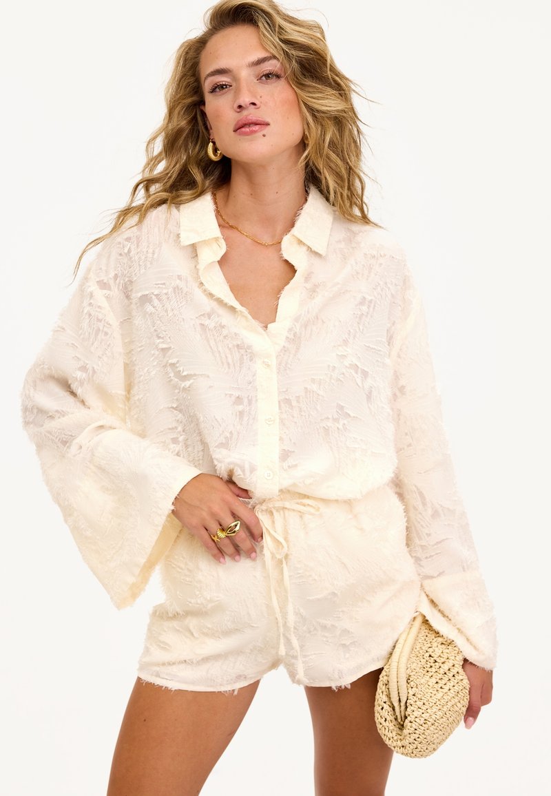 Femme portant une blouse crème texturée à boutons et un short assorti, tenant une pochette en paille tissée, accessoirisée avec des bijoux en or.