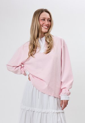 Smilende kvinde med langt blondt hår, iført en løs lyserød langærmet top og et hvidt lagdelt skørt, poserende med den ene hånd på hoften.