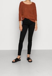 Zrezavě oranžový svetr s texturou a širokým výstřihem, kombinovaný s úzkými černými džíny a černými sandály na plochém podrážku. Jednoduchý a moderní outfit.