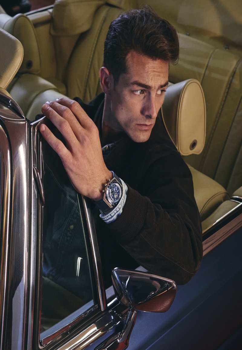 Young man wearing a wristwatch leans out of a vintage car window, looking to his left with a serious expression.