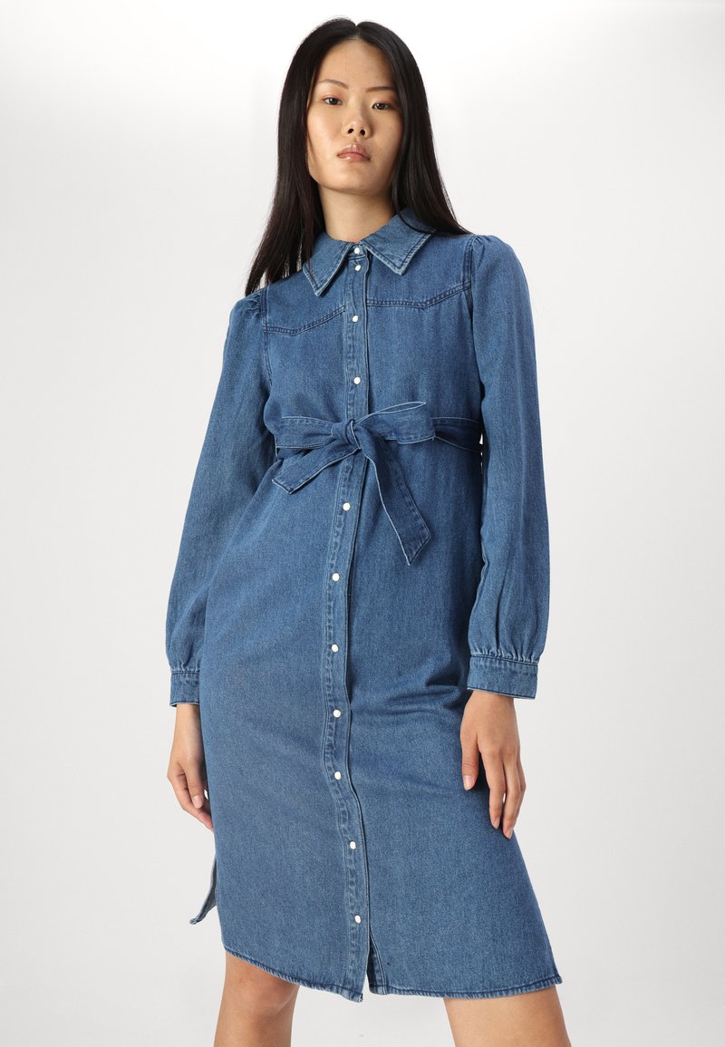 Denim jurkjurkje in medium blauw, lange mouwen met knoopmanchetten, kraag en een strikriem in de taille. Heeft een knoopsluiting aan de voorkant en zijsplitsen.