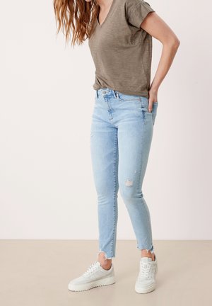 Lichtblauwe skinny jeans met distressed details en rafelige zomen, gecombineerd met een effen olijfgroen V-hals shirt en witte sneakers.