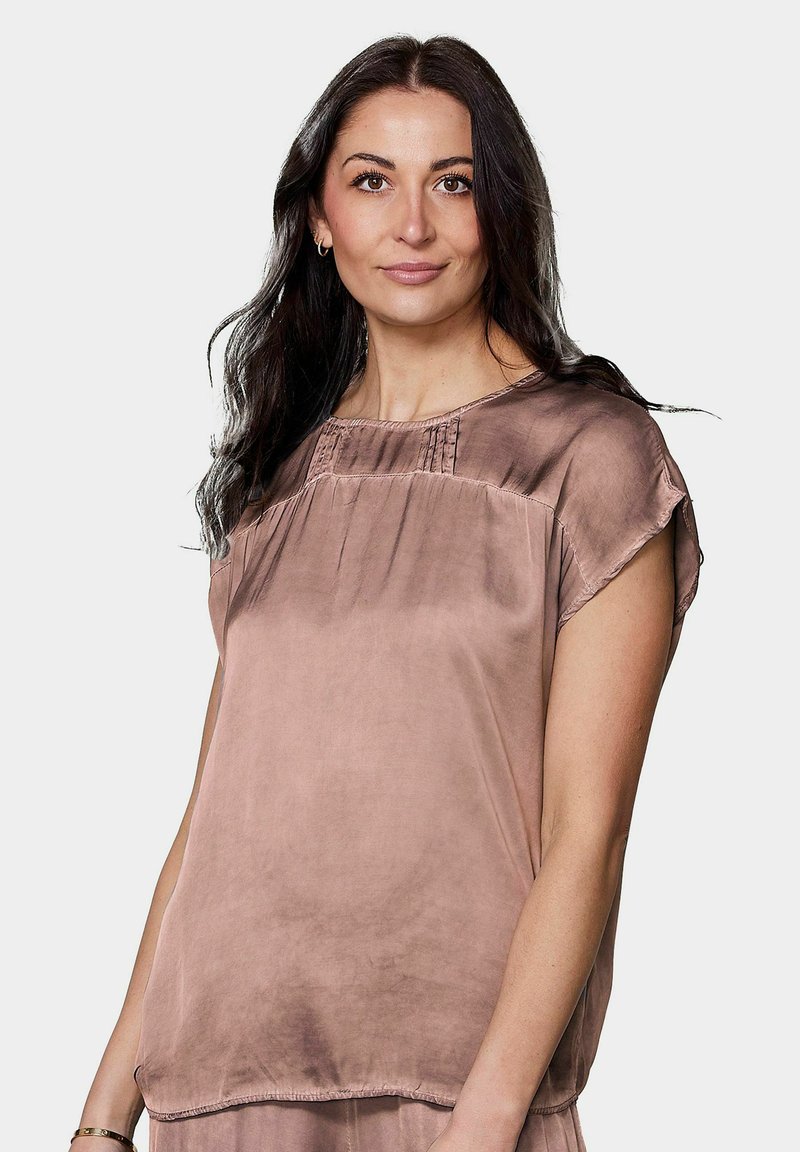Femme aux longs cheveux foncés portant un chemisier ample en satin marron clair à manches courtes, regardant vers l’avant avec une expression neutre.