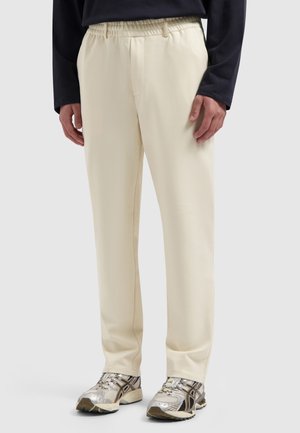 Mann trägt cremefarbene, gerade geschnittene Hosen mit elastischem Bund und silber-schwarzen Laufschuhen, steht vor neutralem Hintergrund.