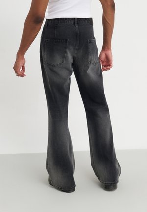 Personne portant un jean noir délavé à jambes larges et un haut blanc sans manches, debout devant un fond clair uni.