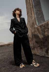 Blazer nero doppiopetto abbinato a pantaloni neri a gamba larga e sneaker con motivo che presentano accenti beige, marrone e nero.