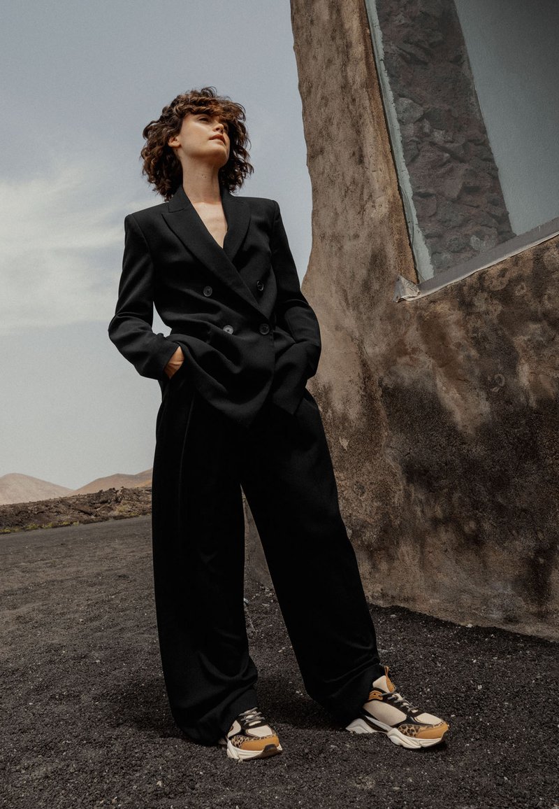 Blazer nero doppiopetto abbinato a pantaloni neri a gamba larga e sneaker con motivo che presentano accenti beige, marrone e nero.