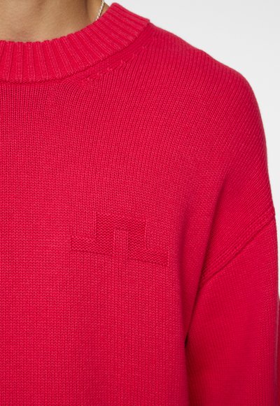 Strikket sweater i klar pink med ribbet halsudskæring og subtilt prægede logo detaljer på brystet. Blød tekstur og afslappet pasform.