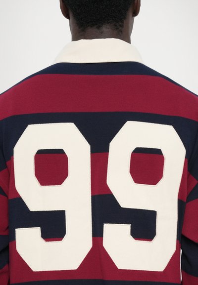 Rugbyshirt in marineblauw met rode strepen; heeft een witte "99" geborduurd op de rug. Zachte textuur met een klassiek kraagontwerp.