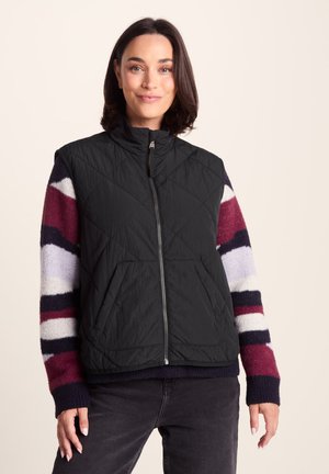 Woman with short dark hair wearing a black quilted vest over a striped sweater with burgundy, white, and navy sleeves, and dark jeans.